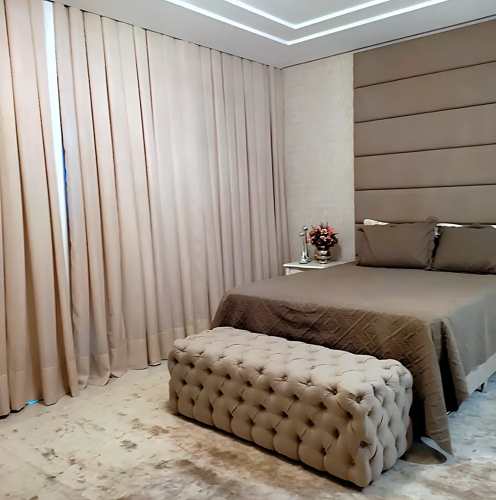 Cabeceira estofada bege em painéis horizontais do chão ao teto, com puff capitonê, em quarto de casal de alto padrão em Salvador